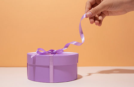 Una mano desata la cinta lila de una caja de regalo redonda sobre fondo naranja