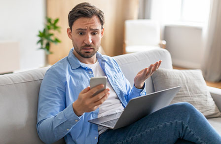Homme perplexe sur un canapé avec téléphone et ordinateur portable